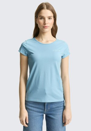 Kobieta stojąca, ubrana w jasnoniebieską koszulkę z krótkim rękawem z drobnym, białym napisem na piersi oraz niebieskie dżinsy, neutralne tło.