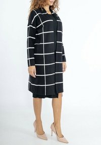Cardigan noir avec un motif à carreaux blancs, manches longues, ouvert sur le devant, en maille texturée, longueur genoux, associé à une robe noire et des talons nus.