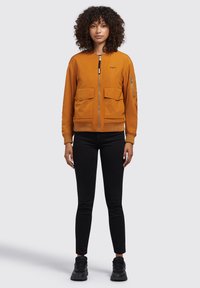 Orangene Bomberjacke mit zwei vorderen aufgesetzten Taschen und einer seitlichen Reißverschlusstasche. Gerippte Bündchen und Saum, Reißverschluss vorne, glatte Oberfläche.
