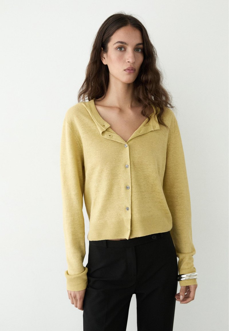 Femme aux cheveux bruns ondulés portant un cardigan boutonné jaune clair à col ouvert et un pantalon noir, debout devant un fond clair uni.