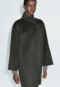 Osoba v tmavě šedém vlněném kabátu s vysokým límcem a širokými rukávy, oblečená nad světle šedým tričkem s dlouhým rukávem, stojící proti jednobarevnému světlému pozadí.