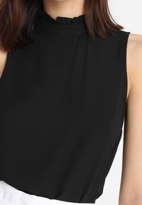 Svart ärmlös topp med en hög volangkrage, tillverkad av ett slätt tyg, har en avslappnad passform och minimala sömnadsdetaljer.
