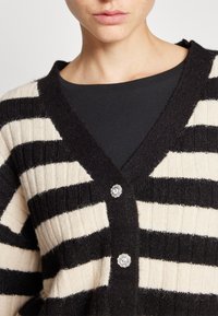 Czarny i kremowy kardigan w paski, z dekoltem w serek, o prążkowanej fakturze i ozdobnymi guzikami z kwiatowym wzorem.