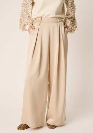 Pantaloni bej din satin, cu talie înaltă și picioare largi, cu buzunare laterale, purtați cu un top texturat cu mâneci lungi și imprimeu și încălțăminte plată împodobită.