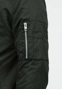 Nærbilde av en svart jakkeærme med vertikal sølvglidelås og rektangulær sydd lomme på ytterarmen.