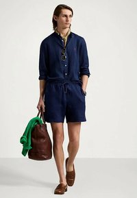 Νεαρός άνδρας φορώντας ναυτικό μπλε πουκάμισο και σορτς, καφέ λοφούς, κρατώντας καφέ δερμάτινη τσάντα με πράσινο πουλόβερ απλωμένο πάνω της, βάδίζει μπροστά.