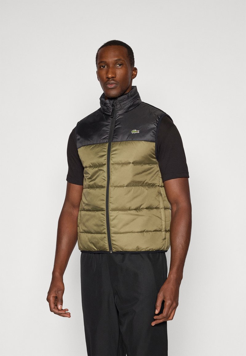 Padded prsluk s gornjim dijelom u crnoj boji i donjim dijelom u maslinasto zelenoj boji. Visoki ovratnik, detalji vertikalnih šavova i Lacoste logo na prsima.