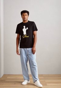 Crni pamučni majica s bijelom grafikom lika koji drži piće, uz tekst "The Simpsons" u žutoj boji ispod. Uparena s svijetloplavim trapericama.