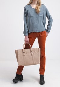 Bolso tote de cuero beige con asas dobles y un logotipo en la parte frontal. El modelo lleva una camisa de botones azul claro y pantalones de color óxido con botas de plataforma negras.