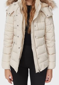 Puffet beige jakke med en blød pelsforet hætte, trykknap- og lynlås-lukninger samt horisontelt quiltet design. Metal detaljer på lommerne.