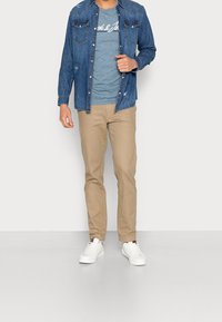 Man in een denim overhemd over een blauwe graphic tee, beige broek en witte sneakers, staande tegen een effen lichte achtergrond.