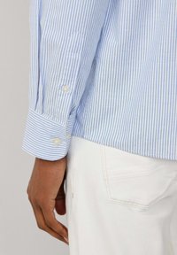 Camicia a maniche lunghe blu chiaro e bianca a righe con polsini abbottonati, realizzata in tessuto liscio. Mano visibile appoggiata su pantaloni bianchi.