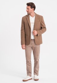 Bruine blazer met notch-lapels, enkele borstzak, gecombineerd met een witte polo en lichtbruine corduroy broek. Afgemaakt met witte sneakers.
