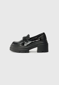 Mocassins noirs en cuir verni avec une semelle plateforme noire épaisse, dotés d'une chaîne décorative à l'avant. Texture lisse, nez arrondi.