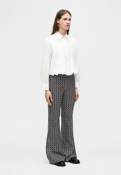 Femme debout portant un chemisier blanc à manches longues avec col et un pantalon noir ample à motif géométrique circulaire blanc et beige.