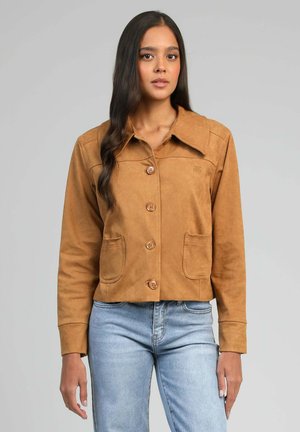 Jeune femme aux longs cheveux foncés portant une veste marron boutonnée avec un grand col et des poches avant, associée à un jean bleu clair.