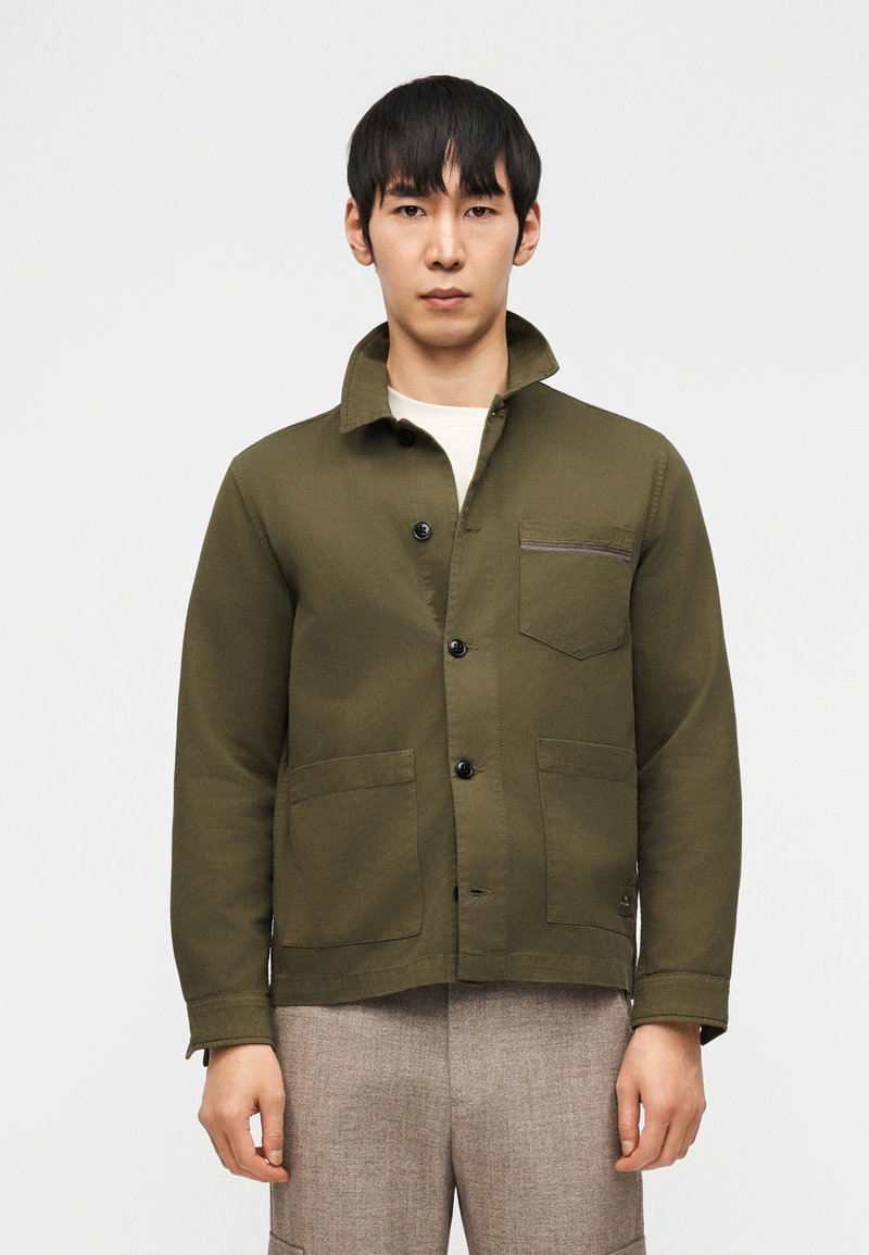 Άνδρας που φοράει χακί πράσινο σακάκι με κουμπιά και τσέπες μπροστά, και μπεζ παντελόνι, στέκεται μπροστά σε απλό λευκό φόντο.