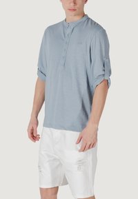 Camicia henley azzurro chiaro con patta a bottoni e maniche arrotolate, abbinata a pantaloni corti in denim bianco strappato con orli sfilacciati.