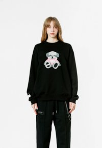 Sudadera negra con un gráfico de oso de peluche en tonos de gris y rosa. La modelo la combinó con pantalones negros sueltos, mostrando un ajuste relajado.