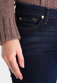 Jeans de mezclilla azul oscuro con herrajes y remaches dorados, que presentan un diseño ajustado y una textura sutil. Mano descansando sobre la cinturilla.