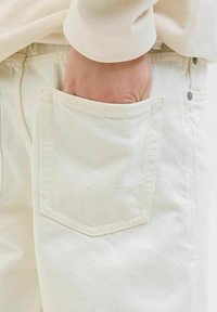 Jeans de mezclilla blanca con un bolsillo trasero con costuras sutiles, complementados por una camisa de manga larga beige clara parcialmente visible.