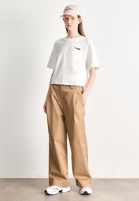 Camiseta blanca recortada con logo verde, combinada con pantalones anchos de tiro alto en color beige. Se lleva con una gorra rosa y zapatillas blancas.