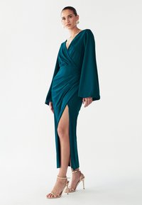 Vestido largo de manga larga en color verde azulado con diseño cruzado, que presenta un escote en v profundo y una abertura lateral. Textura suave con mangas acampanadas. Combinado con zapatos de tacón dorados con tiras.