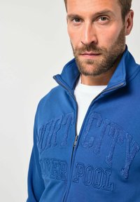 Chaqueta azul con cremallera y texto en relieve "PORT CITY" en el pecho. Textura suave, cuello alto y una camiseta blanca debajo.