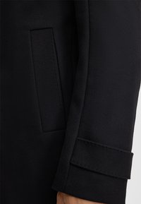 Schwarze Mantelärmel mit rechteckiger Tasche und genähtem Bündchen-Detail am Arm einer Person vor einem hellen Hintergrund.