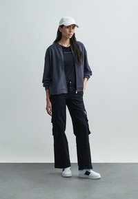 Pilka bomber striukė su užtrauktuku, juoda marškinėliai, juodos krovininės kelnės, balti sportbačiai ir šviesiai pilka kepurė, ant paprasto fono.