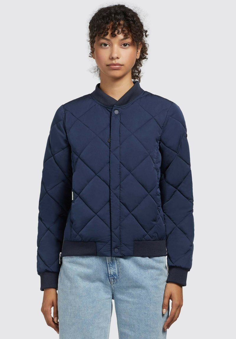 Chaqueta bomber acolchada en color navy con cuello redondo, cierre de botones a presión y puños de canalé. Tejido texturizado con un patrón de diamantes.