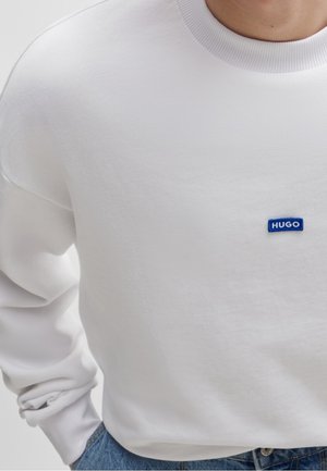 Weißes Sweatshirt mit langen Ärmeln und kleinem blauem "HUGO"-Logo auf der Brust, getragen in blauen Jeans mit hohem Bund.