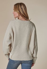 Cremefarbener Pullover mit weichem, strukturiertem Strick, weiten Ärmeln und einem gerippten Saum. Auffälliges Etikett am unteren linken Rücken.