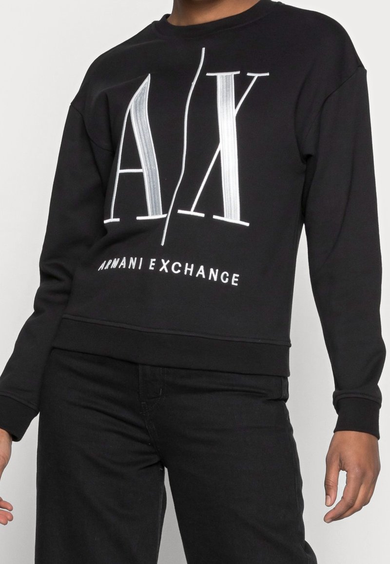 Personne portant un sweat-shirt noir Armani Exchange avec de grandes lettres blanches "A|X" et un pantalon noir assorti, les mains détendues le long du corps.