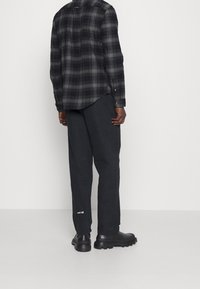 Black plaid button-up shirt with a curved hem, paired with dark gray trousers. Black chunky boots, featuring a white logo on the pants.