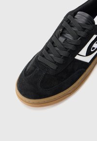 Black suede sneaker with black laces and a white logo accent. Features a rounded toe and a gum rubber outsole.