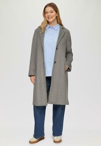 Lichtgrijze wollen jas met inkepingen op de kraag, een enkele rij knopen aan de voorkant en zijzakken, gedragen over een lichtblouse en blauwe spijkerbroek.