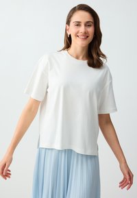 Femme souriante aux cheveux bruns mi-longs portant un t-shirt blanc à manches courtes et une jupe plissée bleu clair sur un fond uni.