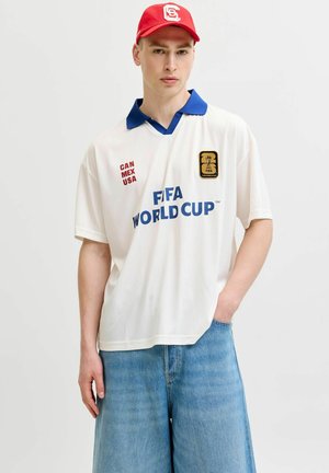 Jeune homme portant un maillot blanc de la Coupe du monde FIFA avec un col bleu, une casquette rouge et un jean bleu large, debout avec une main dans la poche.
