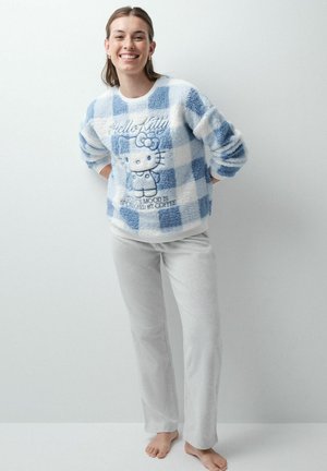 Jeune femme souriante, mains derrière le dos, portant un pull duveteux à carreaux bleu et blanc avec un motif Hello Kitty et un pantalon gris clair, pieds nus.