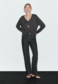 Donkergrijze, oversized long-sleeve top gecombineerd met rechte zwarte denim jeans. Het model draagt minimalistische sandalen en een eenvoudige hangersierraad.