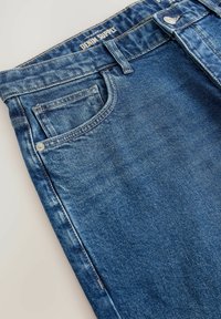 Denim džinsi vidēji zilā krāsā, ar priekšējo kabatu, nolietotu tekstūru un redzamām šuvēm. Zīmola etiketē uz jostas ir rakstīts "Denim Supply".