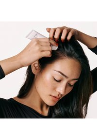 Olaplex haarbehandeling in een witte tube, vastgehouden door een persoon met lang, donker haar. De persoon draagt een zwarte top en heeft een gladde huid met sproeten.