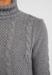 Pull gris à col roulé en maille avec des motifs détaillés de torsades et des points texturés sur la manche et le panneau avant.