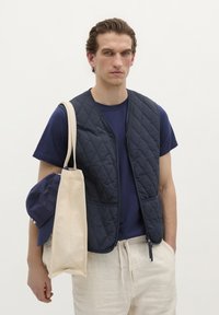 Vesta matlasată de culoare bleumarin cu fermoar, purtată peste un tricou bleumarin. Modelul ține o geantă de umăr crem și o șapcă de baseball bleumarin.