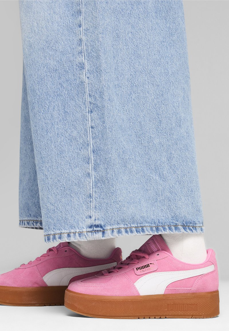 Sneakers in suede rosa con dettagli bianchi e suola in gomma marrone, indossate con jeans a gamba larga blu chiara e calze bianche.