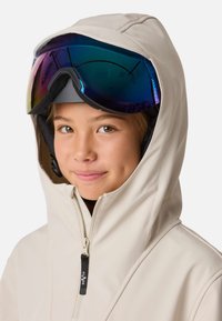 Giacca beige con cappuccio e collo alto, dotata di cerniera frontale e dettagli neri. Il modello indossa occhiali da sci con lente specchiata.