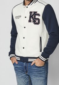 Veste universitaire pour hommes avec un corps crème et des manches bleu marine, dotée d'un grand logo "KS", de poignets côtelés et de poches latérales.