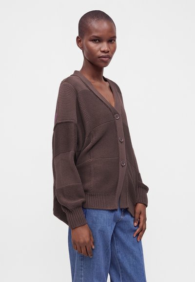 Femme portant un cardigan en maille marron ample boutonné et un jean bleu, debout sur un fond clair uni.