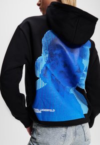 Svart hooded sweatshirt med en blå grafisk design som har ränder och en silhuett på baksidan. Logotyptexten är tryckt i vitt vid fållen.